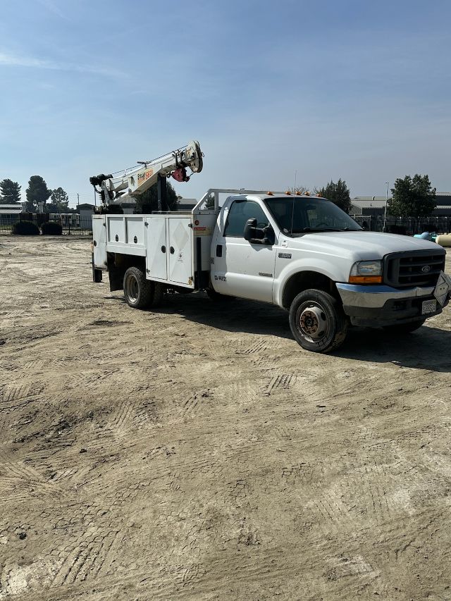 Ford F-550 utility truck with crane, white body, side compartments, on dirt lot.