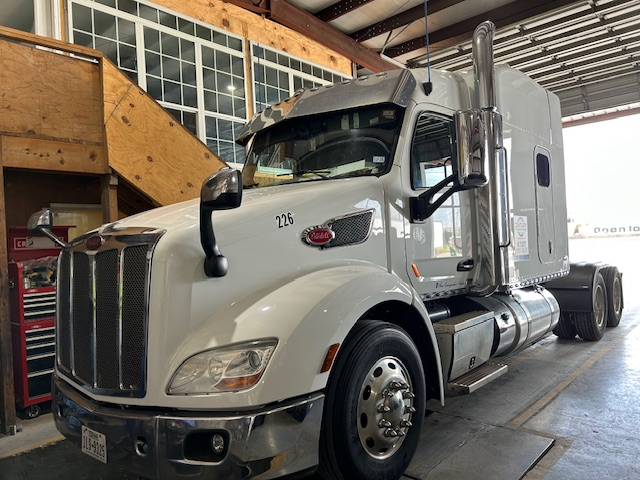 Peterbilt 579 conventional sleeper truck in light gray, number 226 on door, chrome grille, in-service with twin rear axles, inside shop.