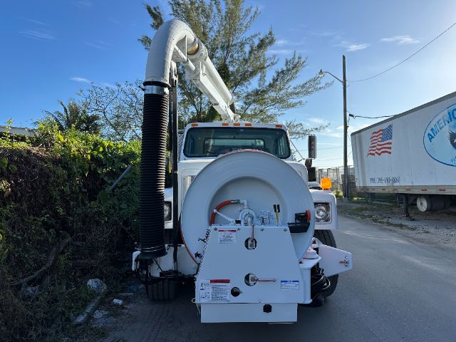 Front view of a white vacuum truck with a large circular hose reel and robotic arm. Visible labels on the rear equipment indicate technical specifications and safety warnings.