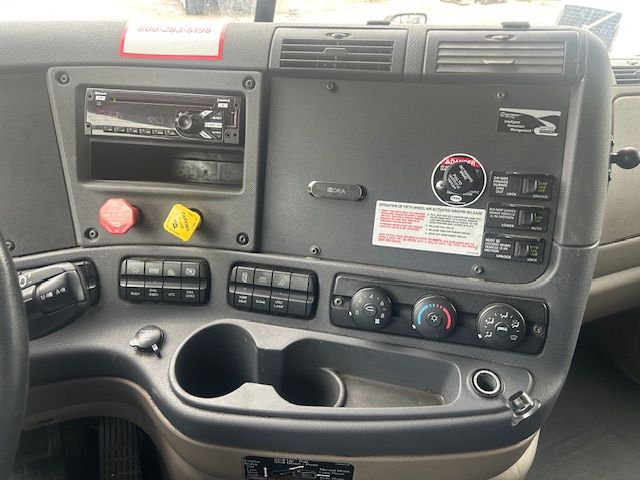 Cabin dashboard of 2019 Freightliner Cascadia 125 showing multimedia stereo, control switches, climate controls, storage cups, and warning stickers.