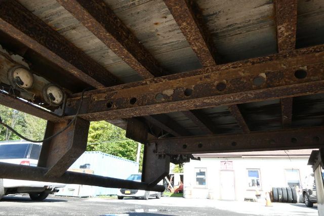Fruehauf FB9T248 trailer chassis showing rusty steel I-beam frame with riveted holes, underside view, supporting cross members, and some vehicles in background.