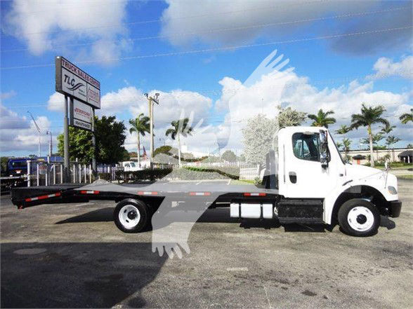 Flatbed truck side view showing no visible branding text on cab; long flatbed with wheelbase and mirrors; background shows palm trees and dealership signage.