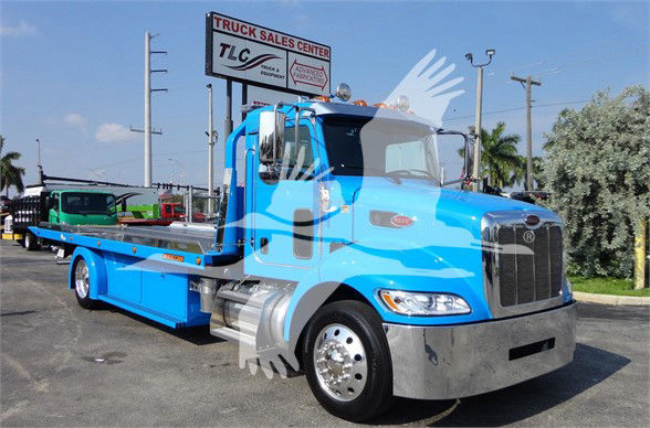 Blue Peterbilt 337 tow truck with flatbed seen front-right, dealership sign in background.