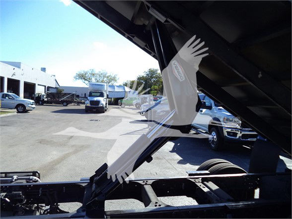 Isuzu dump truck with hydraulic bed raised, visible cab and engine area; background includes other trucks in lot.