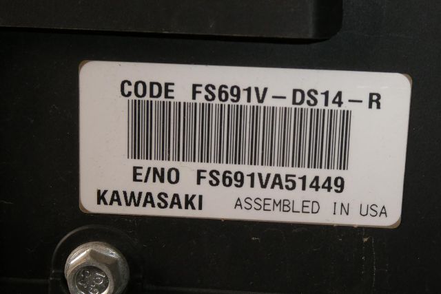 Code sticker on equipment showing FS691V-DS14-R, E/NO FS691VA51449, KAWASAKI, ASSEMBLED IN USA, barcode, and screw in foreground.
