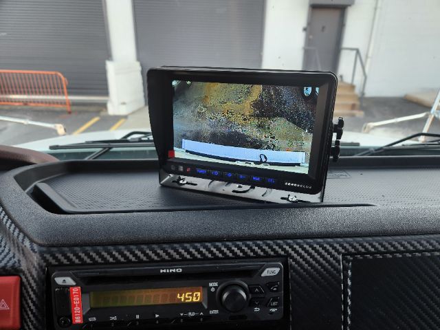 Dashboard of a 2025 Hino L6 truck interior with a mounted tablet screen showing a rear view camera feed; Hino radio/navigation unit visible; carbon fiber style dash trim.