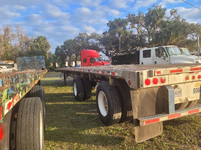 Great Dane flatbed trailer with dual rear axles and a metallic deck. Visible rear lights, mud flaps, and a license plate area. Surrounding trucks and a blue sky with clouds.