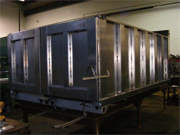 Dump body chassis on a trailer with metal side panels and vertical reinforcing ribs. Visible: weathered metal with riveted seams and latch; no text readable on panels.