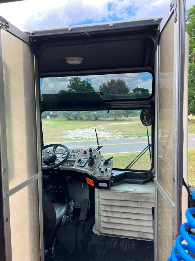 Industrial truck cab interior with steering wheel, control panel, pedals, and a white ribbed equipment housing; open doors with a view to outside fields.