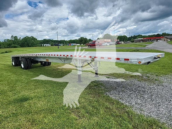 Flatbed trailer on grassy lot showing long aluminum deck with tandem axles and reflective safety stripes.