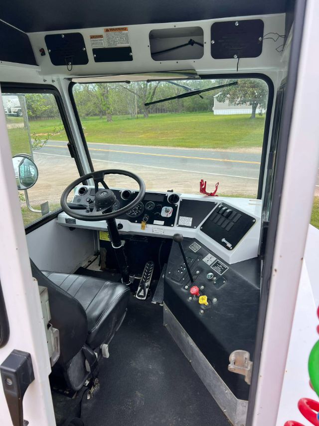 Truck cab interior showing steering wheel, dashboard, pedals, and control levers in a yard truck. Windows reveal exterior road and greenery.