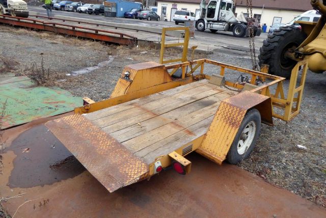 Trailer deck with wooden planks, rusted metal fenders, and a rear loading ramp. Industrial yard in background with heavy equipment.