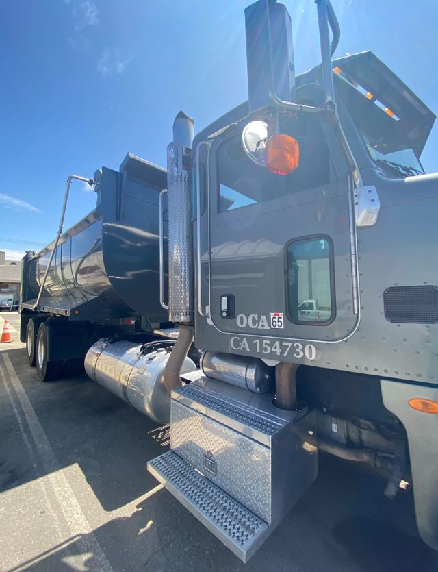 Peterbilt 365 dump truck rear cab area with chrome chrome steps, exhaust stack, and tailgate raised. Visible CA license plate number OCA65 and CA154730.
