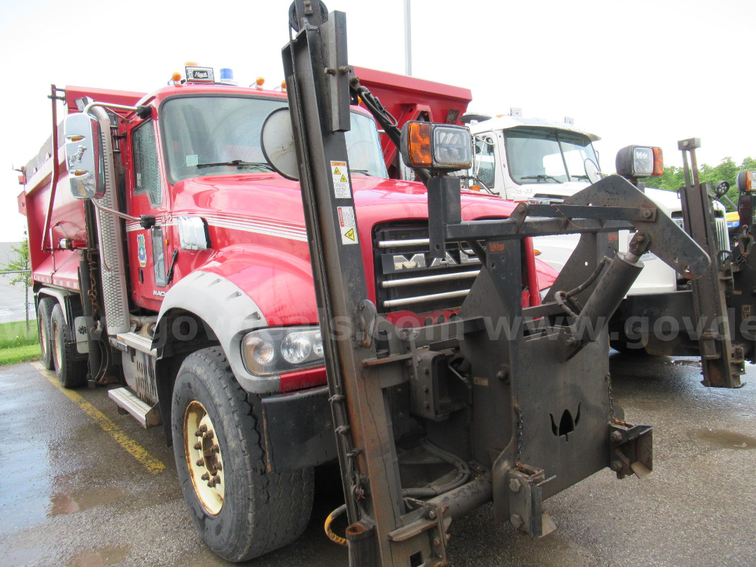 Rear/front view of a red 2014 Mack GU713 plow truck with a large mechanical attachment in the foreground.