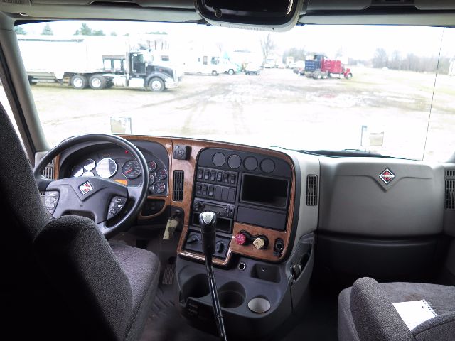 Interior of a 2010 International PROSTAR cab showing steering wheel, dashboard with wood trim, gear shifter, and center console; windshield view of a truck yard.