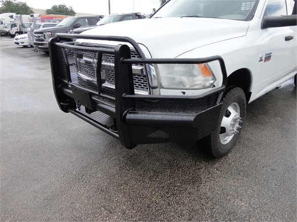 Ram 3500 pickup with heavy-duty black grille guard and bumper at front showing chrome headlamps and badge. White cab, front wheel visible, rain-soaked lot.