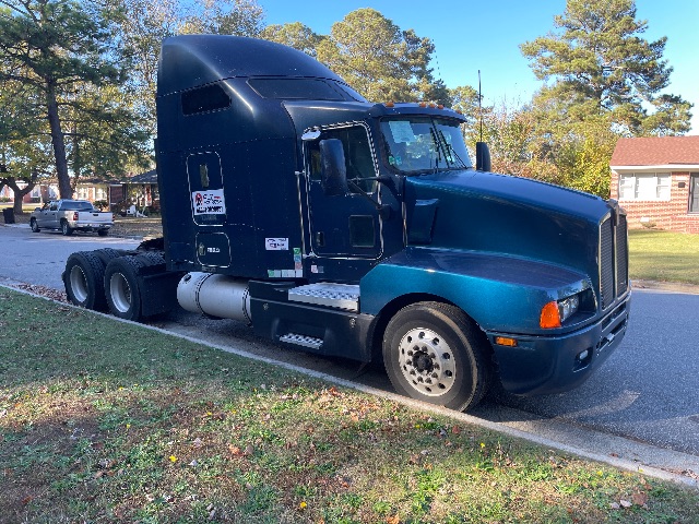 Kenworth T600 tractor unit in dark teal, side view shows sleeper cab, dual rear axles, exhaust and fuel tanks visible. No legible labeling readable on image.