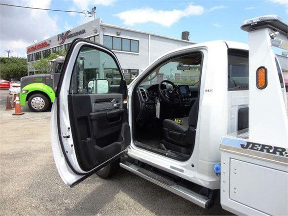 Ram pickup truck interior visible through open driver door; black interior, steering wheel, dashboard, and pedals. Background shows a dealership with other trucks and a green vehicle.