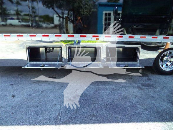 Tow truck with chrome bumper and cutouts, visible side compartments in the lower bumper area; reflection of a person in glass, truck wheel on right.