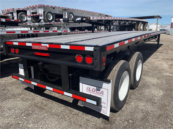 Flatbed trailer showing dual rear axles with white wheels and red taillights; Fontaine brand plate visible; additional trailers and equipment in background.