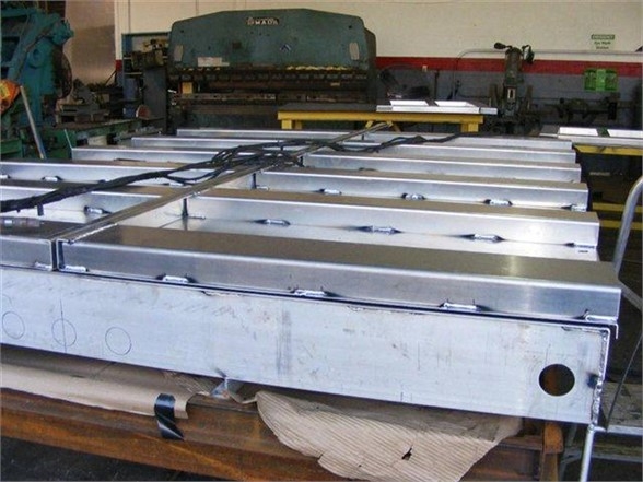 Metal truck bed or dump body components with mounting rails and lugs, aluminum-looking panels, workshop setting.