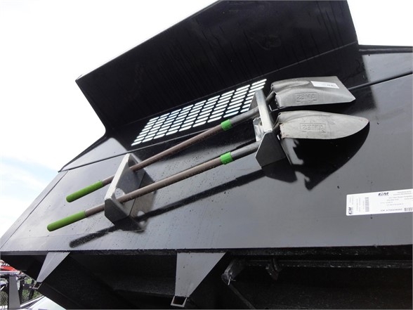 Dump truck tailgate with shovels and tools resting on edge, green handles, metal levers, and a grill vents.