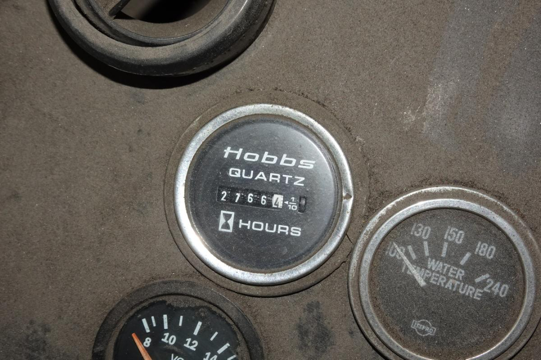 Hobbs Quartz hour meter reading 27,664 hours; gauge cluster with water temperature gauge showing 130-180 degrees; part of dashboard showing dusty surface.
