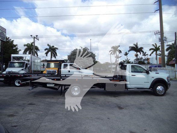 Flatbed tow truck with white cab and black flatbed, parked in lot with other trucks and palm trees in background.
