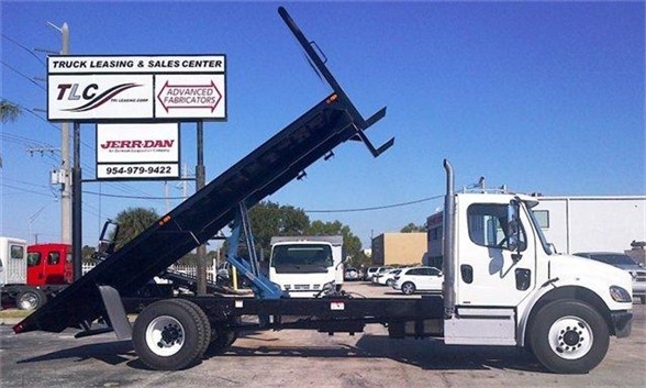 Flatbed tow truck with hydraulic tilting bed raised, white cab, black flatbed, in a truck lot.