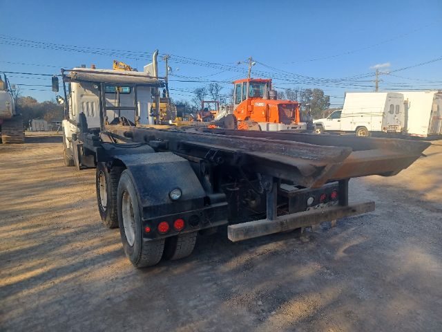Rear view of a Volvo VG64 roll-off truck with large dumping container and dual rear axles, in a yard with other trucks. No legible labels visible on components.