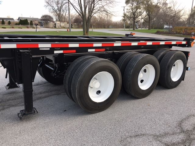 Trailer chassis showing triple-axle wheel arrangement with black frame and white rims, side rails with red and white reflective strips.