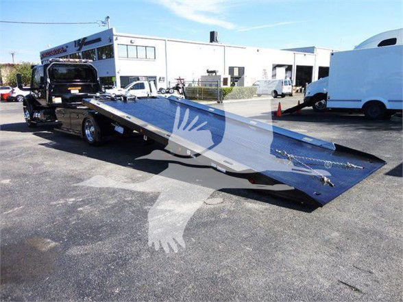 Tow truck flatbed trailer on a service lot with a ramp extended for loading, showing a white building backdrop and several vehicles.