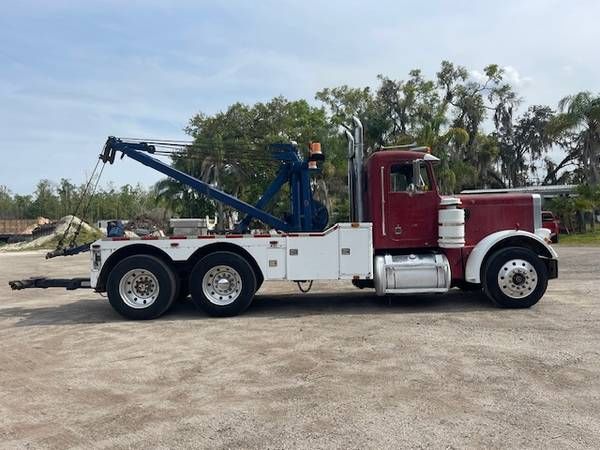 Peterbilt 359 tow truck with a red cab and white utility bed, wheel-lift apparatus extended on a dirt lot.