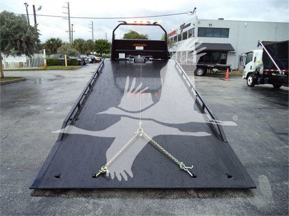 The image shows a black tow flatbed with ramps and railings, with a chain secured at the front. There are markings on the bed including a large white hand and eagle design watermark across the surface.