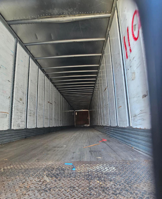 Trailer interior showing long metal box trailer with ribbed walls, wood floor, overhead supports, viewed from the open back.