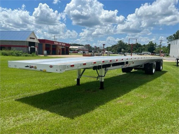 Long aluminum flatbed trailer on grass with dual rear axles, side marker lights, and a support leg visible in foreground. Clear blue sky with clouds.