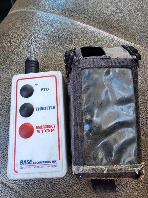 Trucking control remote with two black buttons labeled PTO and THROTTLE and a red Emergency Stop button, placed beside a rugged case containing a cracked screen device.