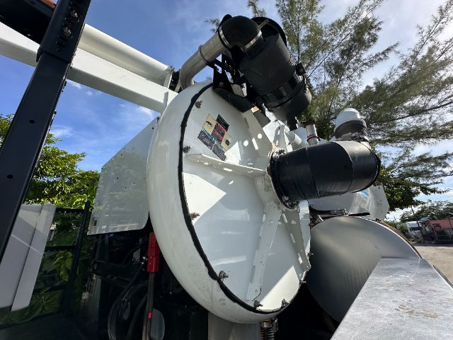 Industrial vacuum truck with large round vacuum chamber and black ducting fittings, white metal housing, blue sky, trees in background.