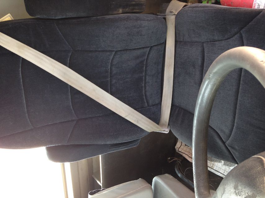 Truck seat in cabin with seat belt across, steering wheel visible at bottom left, dark fabric upholstery.