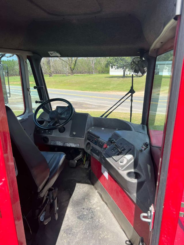 Truck cab interior showing steering wheel, dashboard, two seats, and front window with wiper; fan mounted near roof; red exterior panels visible.