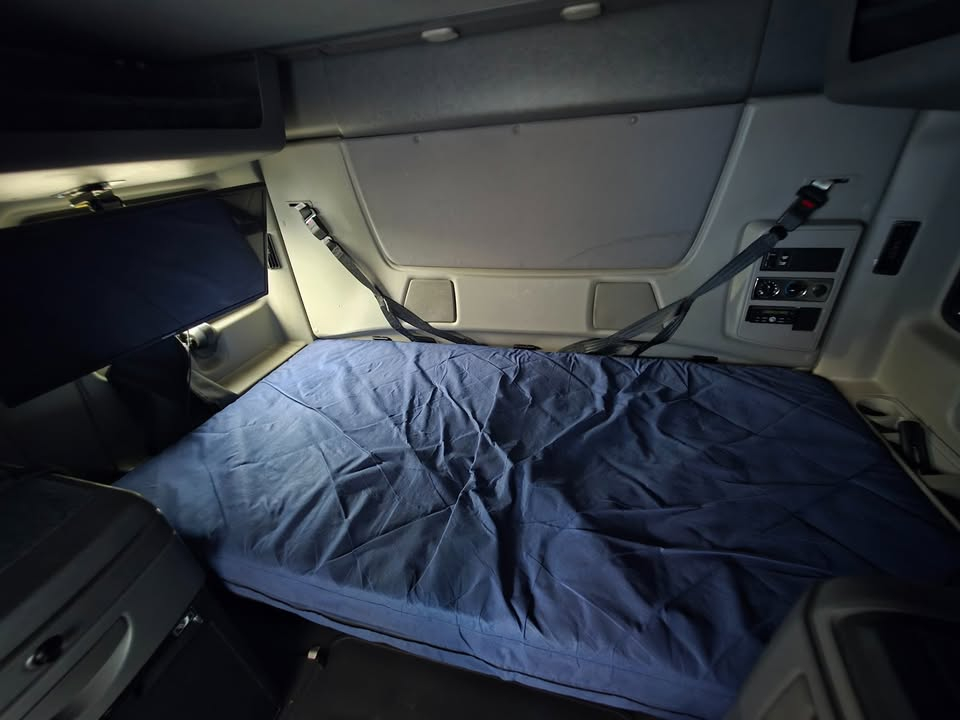 Truck cab interior showing a blue blanket or cover over the sleeper area, side panels with controls, and a window panel.