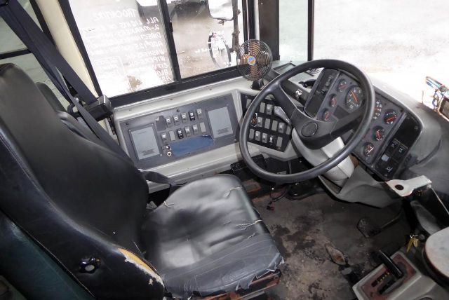 Bus interior driver's area with steering wheel, instrument cluster, and control panel. Worn driver seat, large windshield, dashboard with gauges and switches.