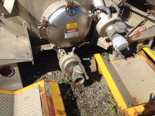 Tank vehicle rear showing circular vacuum tank with hoses and valves, warning labels, and metal steps/grated platform.