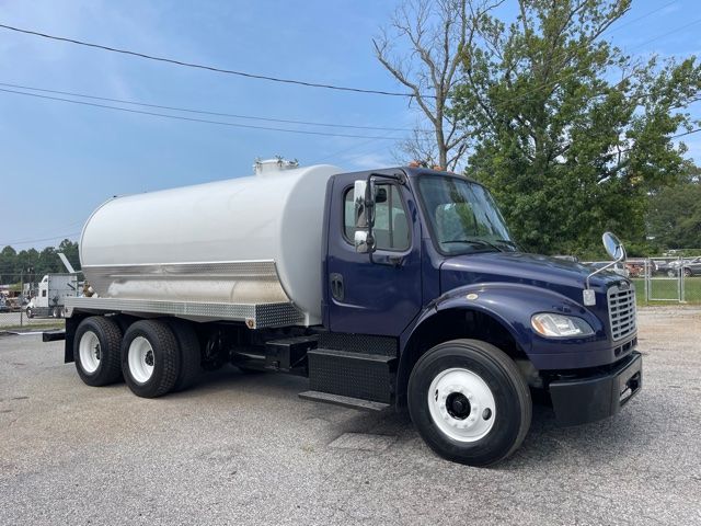 Truck showing a 2013 Freightliner M2 sewer service with a large cylindrical stainless steel tank mounted on a dark blue cab, six wheels, side step, and front bumper.