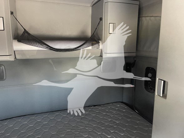 Kenworth T880 sleeper cab interior showing storage compartments and a netted shelf above a padded sleeper mattress; side wall and cabinet door visible.