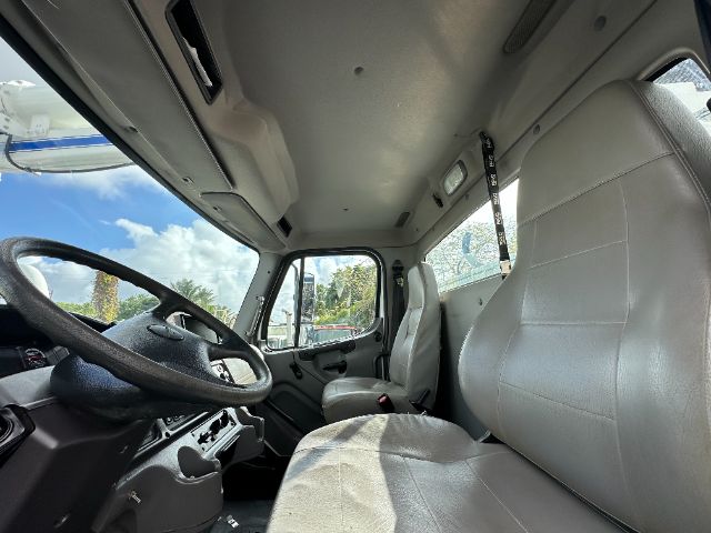 Truck cab interior showing steering wheel, driver seat, and passenger seat with light gray upholstery; dashboard with controls; large windshield and side window; overhead storage area.