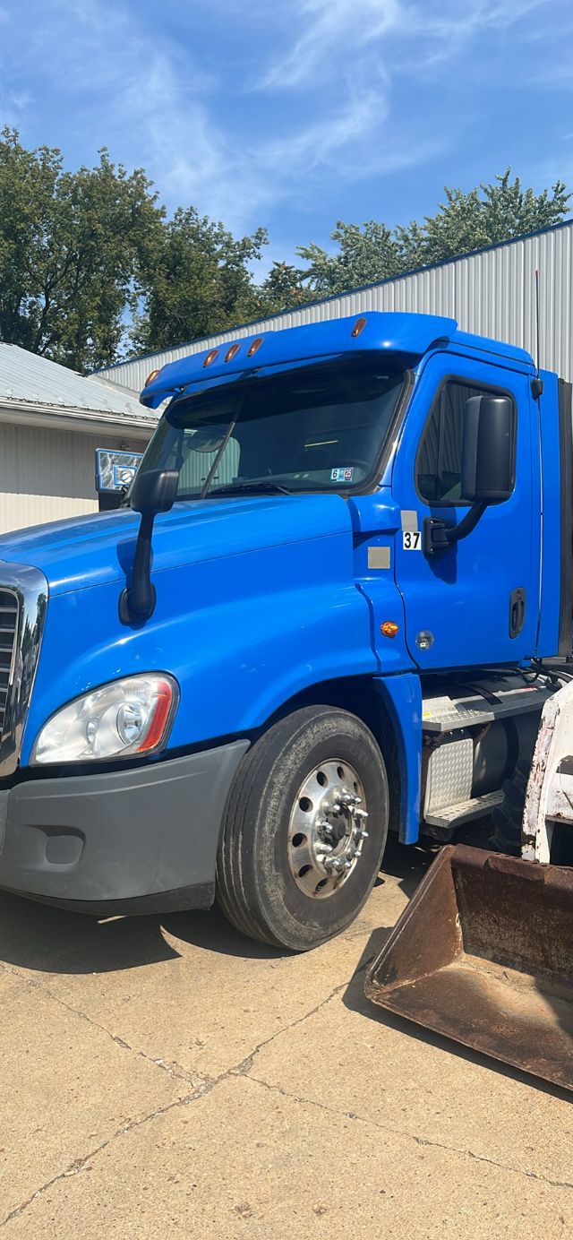 Freightliner Cascadia truck cab in blue with side mirror and windshield, front wheel, and a metal bucket attachment in foreground.