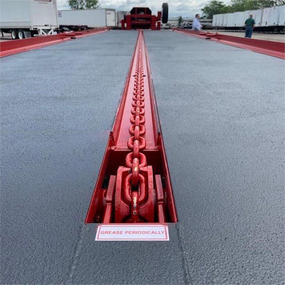 Long red drop deck container delivery trailer with chain and coupler, grease periodically warning label visible, on a flatbed surface.