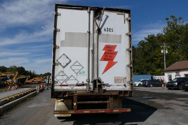 Rear view of a white cargo trailer with two doors, various diamond placards, red warning arrow, and numbers 6385 on the door. Behind are other parked vehicles and yard equipment.