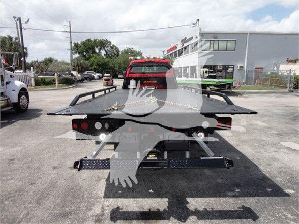 Tow truck flatbed rear view with heavy-duty steel ramp, chain, and taillights visible. The image shows a red vehicle on a service lot in front of a gray building.
Visible text includes: none clearly legible from the image
